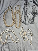 Top view of 6 pearl necklaces in cream, white, and gray-silver colors arranged on a gray fabric background.