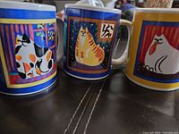 Overview photo showing set of three ceramic mugs with colorful cat artwork, each with different design.
