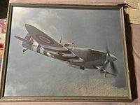 Front view of framed Spitfire plane art print showing aircraft flying against cloudy sky.