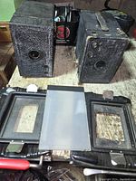 Two vintage Cartridge Hawkeye Model B box cameras and a photographic processing plate holder with two framed plates and one translucent plate, placed on a wooden surface.
