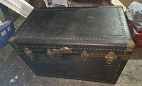 Front angled view of black vintage trunk with metal studs and brass lock and side handles.
