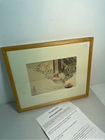 Framed antique Japanese print showing a young child leaning over a platform watching a small fire. Frame is wooden with white matting around the print.