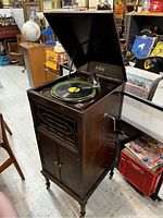 Full view of McLagan phonograph gramophone cabinet in wood with open top lid, showing turntable with a record on it and crank handle on side.