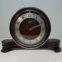 Front view of Art Deco mantel clock showing circular face with walnut center and silver outer ring with numbers. Clock sits on a wide layered wooden base with curved design edges. Chrome and bakelite detailing visible.