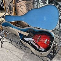 Guitar inside silver hardshell case with blue plush interior, showing redburst top, f-holes and trapeze tailpiece
