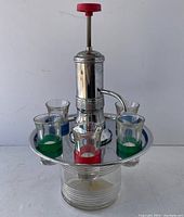 Front view showing liquor dispenser with pump top on glass container and six shot glasses arranged on the chrome tray.
