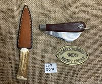 Three items laid on textured burlap: the hawkbill cutter with wooden handle, the antler-handled knife in leather sheath, and oval brass plaque with embossing.