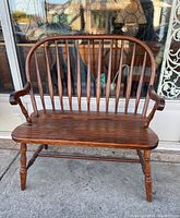 Front view of the oak Windsor style bench showing curved backrest with spindles, armrests, contoured wooden seat, and turned legs.