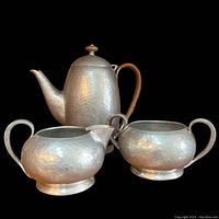 View of three-piece pewter tea set with hammered texture: teapot, creamer, and sugar bowl arranged together against a black background.
