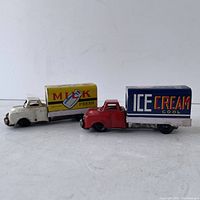 Side view of two vintage tin litho friction toy trucks, Milk Truck in white and yellow, Ice Cream Truck in red and blue, showing overall condition and colors.