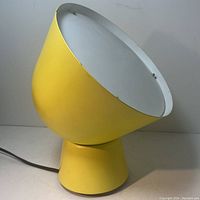 Yellow cone-shaped midcentury lamp with white interior, shown powered with electric cord, placed on surface.