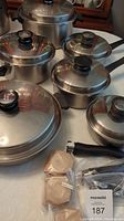 Full set of stainless steel Amway Queen cookware with lids and replacement handles and knobs on table.