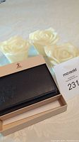 Photo showing the men's black leather trifold wallet in its box, black smooth leather, new in box, placed alongside three rose-shaped candles.