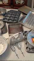 Photo showing metal bakeware including muffin tins, cookie sheets, hand mixer with two beaters, 9x13 pan with insert, Bundt and angel food cake pans, and disposable aluminum pie plates arranged on a table.