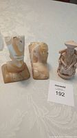 Photo shows three carved sculptures including Nefertiti bust, sphinx, and Asian man figure on a white fabric.