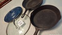 Two cast iron skillets with two lids visible - a blue speckled enamel lid and a glass lid with metal rim and handle.