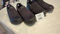 Three pairs of black Skechers slip on shoes arranged on a table with the lot tag visible.