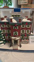 Front view of the Old Chelsea Mansion ceramic Christmas village house in its original styrofoam packaging, showing festive wreath decorations and simulated snow on the roof.