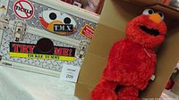 Original metal box packaging next to Tickle Me Elmo doll, showing branding and Try Me button.