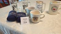Photo showing the collection of various US Navy and Oktoberfest themed mugs and two blue neoprene beverage cozies on a white tablecloth.