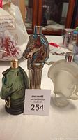 The three Avon cologne bottles displayed on a table with a numbered lot card (254). The bottles include two hitching post shapes in green glass and metallic silver finish, and one frosted glass Centurion helmet shape.