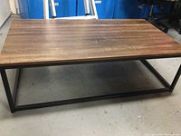 Rectangular coffee table with wood grain top and black metal frame shown on concrete floor with blue bins and other furniture in background.