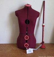 Front view of maroon dress form showing circular adjustment dials and red measuring stick standing adjacent.