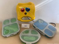 Top view showing the yellow Cookie Monster waffle maker, one green and two blue-tinted compartment dinner dishes with transparent lids.