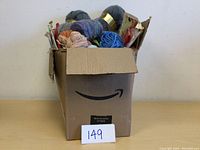 Cardboard box filled with yarn, knitting needles, and patterns from a front angle