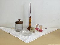 Wide image showing entire lot on white cross-stitched tablecloth, including candy jar with copper lid, amber candlestick with purple candle, small colored glass vases, and glass bowl.