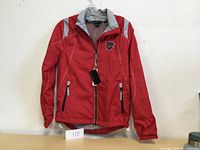 Front view of the red North End windbreaker jacket with a black zipper, gray reflective accents on the shoulders and pockets, and an embroidered logo patch on the chest.