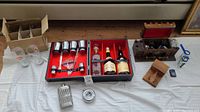Overview of the lot on wooden surface with a box of glasses, two double highball glasses, the barware set with decanters and bottles, silver flask, small round silver item, small wooden box, small green bottle, and other small items.