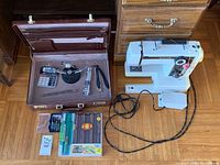 Brown briefcase open showing office supplies inside including calculator, stapler, flashlight, pens, and coin management tools. White Singer sewing machine placed on floor nearby with cords and foot pedal.