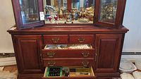 Wide shot of wooden cabinet with drawers open revealing silver plated cutlery in trays and table linens in upper drawer.