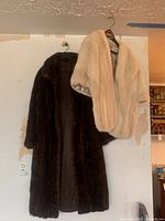 Both garments hanging on wall hooks: the brown fur coat and the white fur shawl side by side.