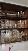 Photo of wooden cabinet shelves with a variety of glassware, etched crystal decanter, metal trays, and metal cups