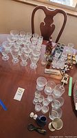 Photo showing assortment of glass stemware including wine glasses, shot glasses, several small decorative bottles, and bar accessories arranged on a wooden table.