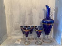 Six cobalt blue wine glasses with gold grape and scroll detailing, shown standing together on a shelf with a price tag visible.