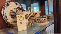 Wide shot of 6 teacups and saucers stacked with 11 dessert plates and creamer in background inside glass cabinet