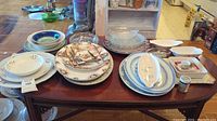 Wide view of assorted porcelain plates, platters, and glass serving bowls displayed on a wooden table.