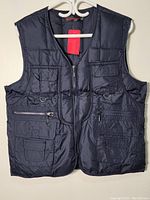 Front view of black sleeveless vest jacket hanging on a white hanger against a wall, showing overall shape and pocket layout.