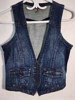 Front view of a dark blue denim vest hanging on a white hanger against a white wall. Shows deep V neckline, front button closure, and two front flap pockets.
