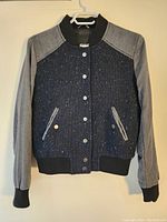 Full front view of blazer hanging on white hanger showing navy textured front, gray sleeves, black ribbed cuffs and hem, silver buttons, and two front pockets.