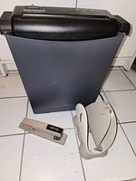 Side view of the paper shredder, beige napkin holder, and beige metal Swingline stapler with staples on the floor.