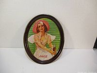 Photo of full oval 1977 Coca Cola serving tray with woman in pink bonnet sitting and holding a Coke bottle against green background.