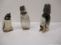 Three carved figures on a plain background. One penguin-shaped figure with black, white, and beige colors; two humanoid carved figures with black and white painted details.