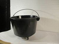 Side view of black cast iron pot with wire handle and three legs on a white surface.