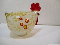 Side view of the glass rooster displaying white opaque glass with yellow speckles and red details including comb, wattles, and circular spots on body.