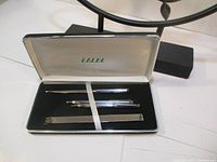 Full view of Lalex pen set box containing two pens, letter opener, and ruler on black velvet interior with white satin lid, items held with elastic band