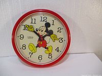 Front view of round red clock featuring Mickey Mouse on the face with black numbers and yellow hands forming Mickey's arms
